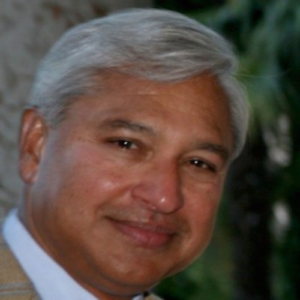 A man with gray hair and a light beard is smiling slightly, wearing a beige jacket. A blurred natural background is visible.