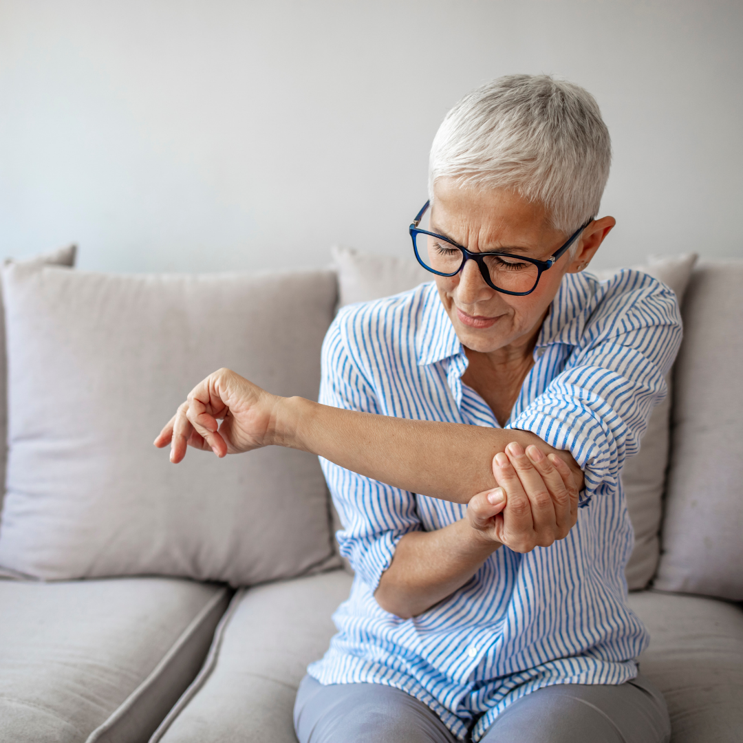 A person with short gray hair and glasses is seated on a couch, holding their right elbow with their left hand, wearing a blue striped shirt.