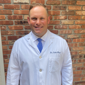 Person in a white lab coat with a blue tie stands in front of a brick wall, smiling at the camera.