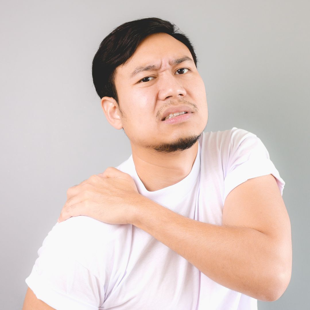 A man in a white t-shirt, possibly contemplating a visit to an orthopedic in Patchogue, holds his right shoulder with a pained expression against a plain background.
